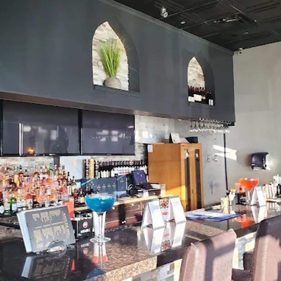 Modern restaurant bar with colorful liquor bottles, turquoise cocktail, marble counter, and upholstered seating beneath arched wall niches.