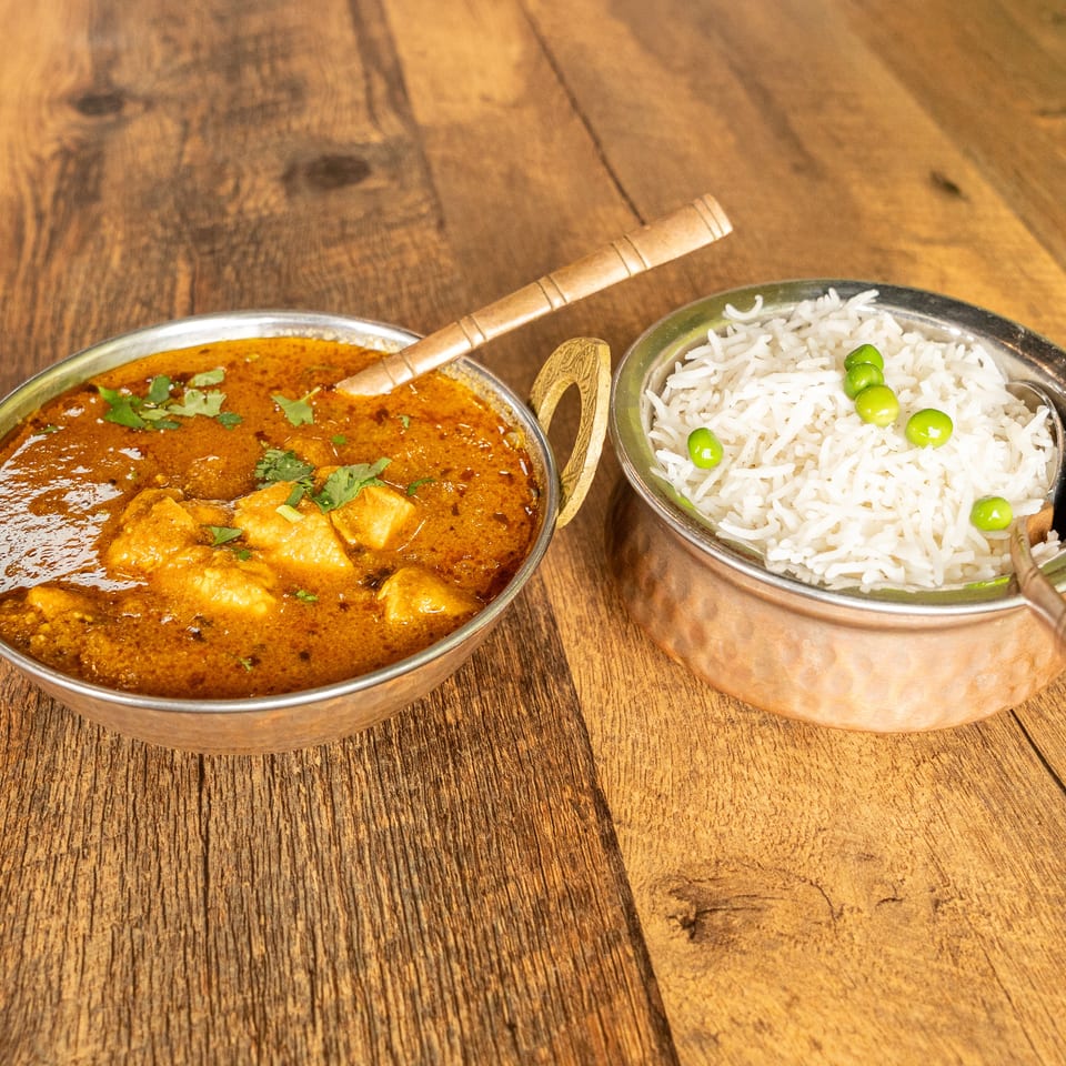 Indian curry garnished with cilantro served beside basmati rice and green peas in metal bowls on a wooden table.