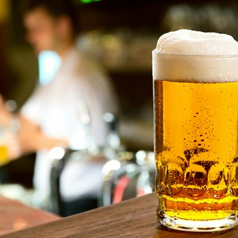 Frosty pint of golden beer on a wooden bar, with a softly blurred bartender and bottles in the background.