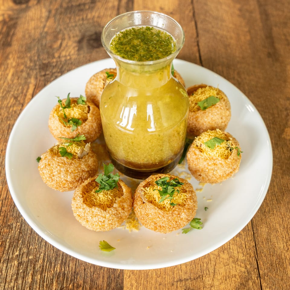 Crispy pani puri shells filled with spiced vermicelli and cilantro surround a glass bottle of tangy green pani on a white plate.