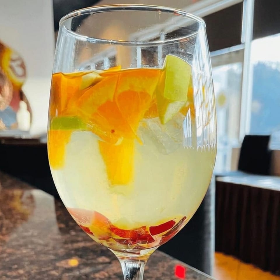 Large stemmed glass filled with pale citrus drink, orange and lime slices, and ice in a sunlit restaurant interior.