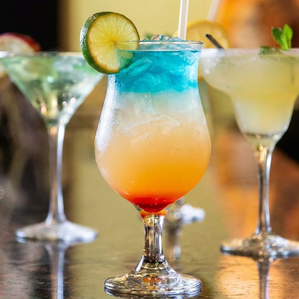 Three colorful cocktails sit on a restaurant table, including a layered blue, orange, and red drink garnished with lime.
