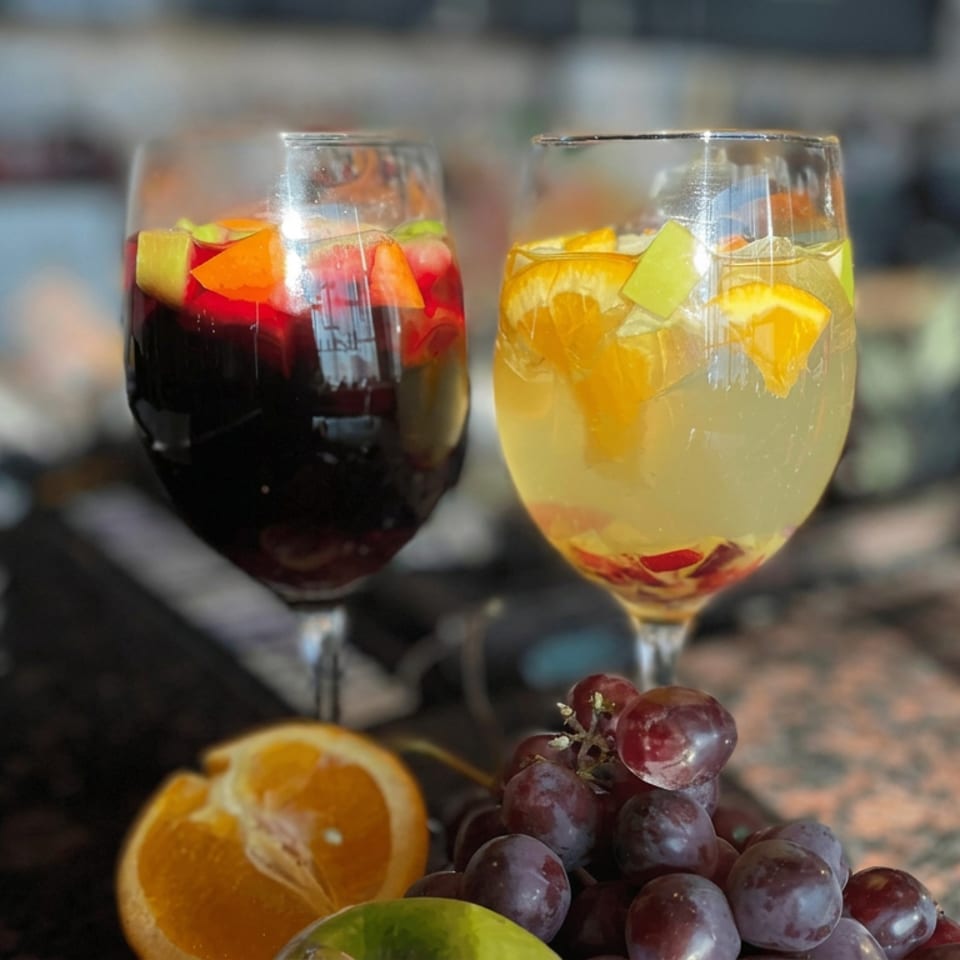 Two glasses of red and white sangria garnished with citrus and fruit on a sunlit restaurant table.